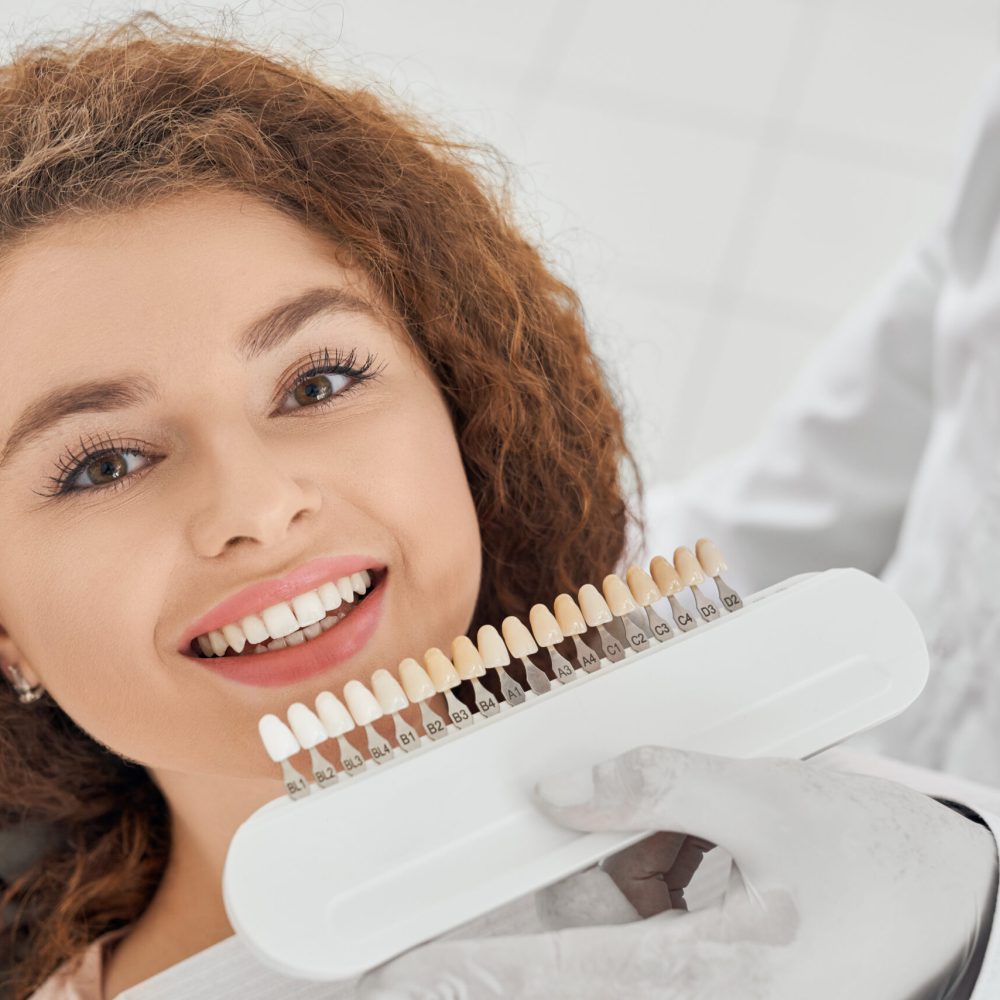 Woman smiling while male dentist keeping teeth color range Closeup of beautiful young woman lying on dental chair, looking at camera and smiling while male dentist keeping in hands teeth color range. Girl doing whitening procedure in dental office.